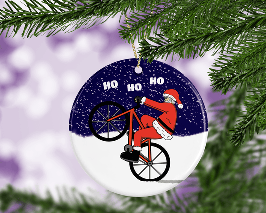 A round ceramic Christmas decoration with an hand-drawn illustration of Santa Claus riding a push bike and pulling a wheelie in the snow.,