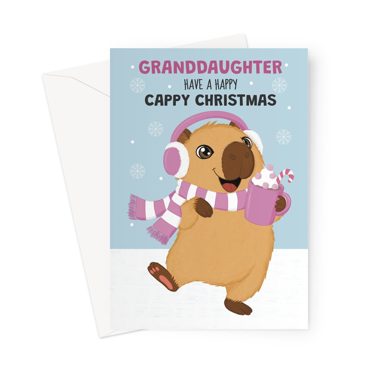 A Christmas greeting card with a white envelope behind it, featuring a cartoon capybara wearing pink headphones and a pink-and-white striped scarf, holding a mug with whipped cream and a candy cane. The card reads 'GRANDAUGHTER HAVE A HAPPY CAPPY CHRISTMAS' on a light blue background with snowflake details.